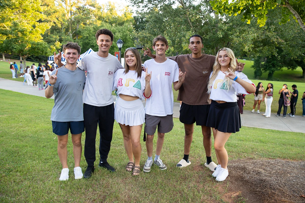 A group of six young people is posing cheerfully outdoors, with some wearing shirts with Greek letters.