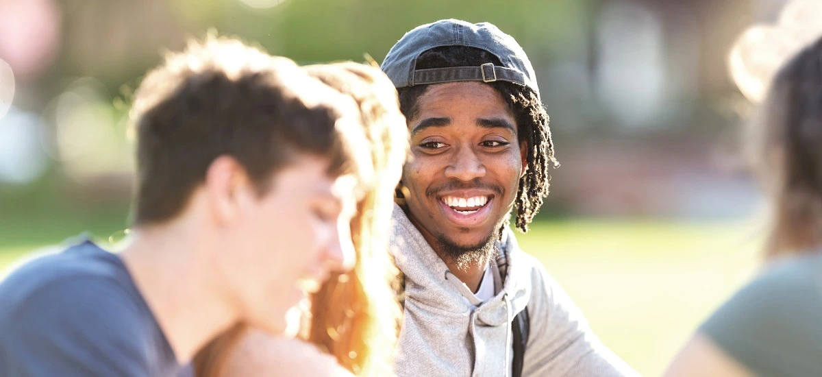 A group of young people are sitting together outdoors, with one person smiling brightly at others.